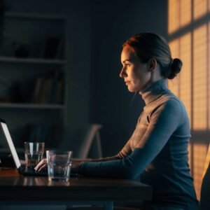 אישה יושבת עם לפטופ וכוס מים ליד חלון שטוף אור, נראית רגועה וממוקדת בעבודה